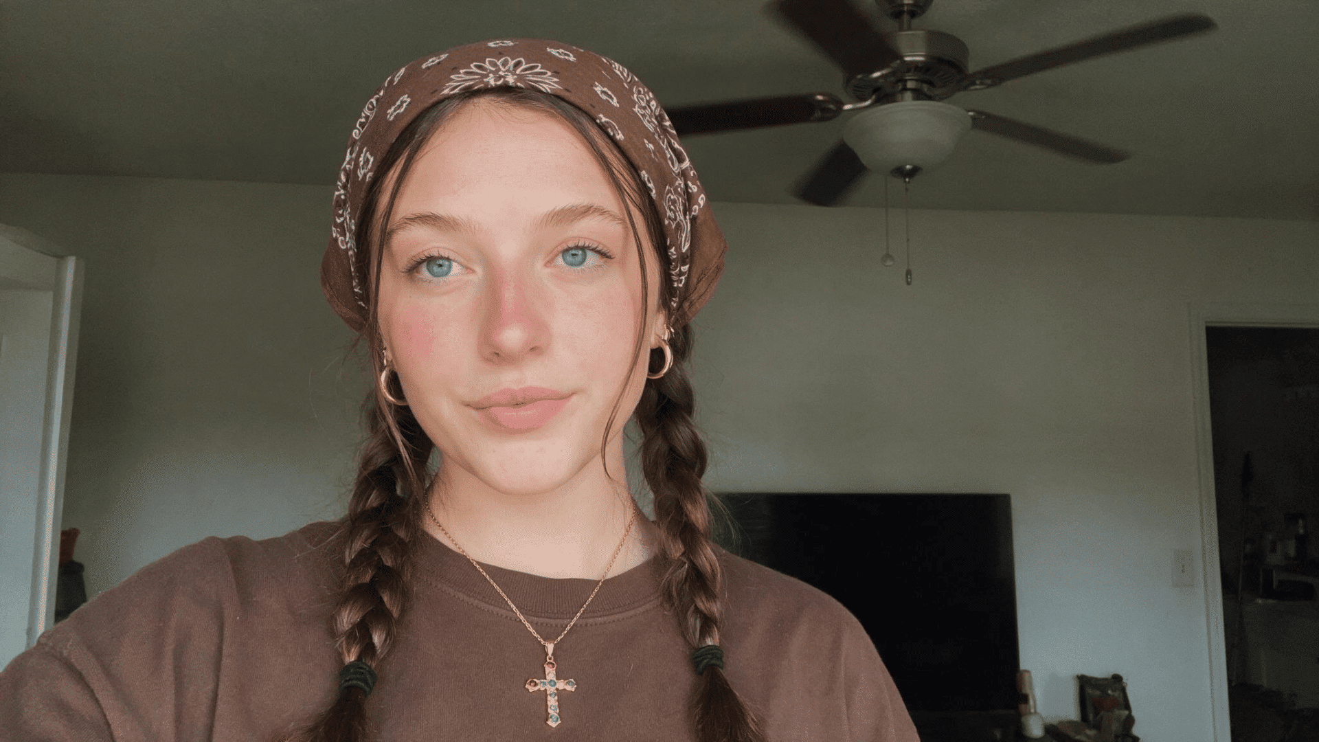 oman wearing a brown paisley bandana with two braids, a brown top, and a cross necklace in a casual indoor setting.