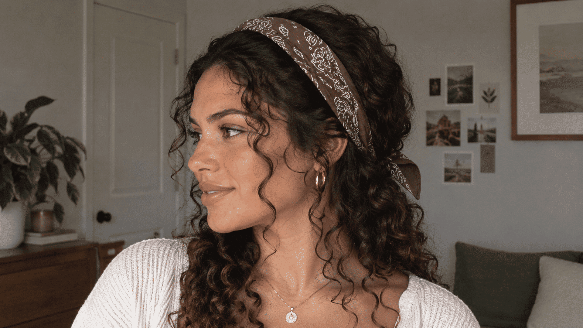 oman with curly dark hair wearing a brown paisley bandana headband, cream top, and gold jewelry in a casual indoor setting.