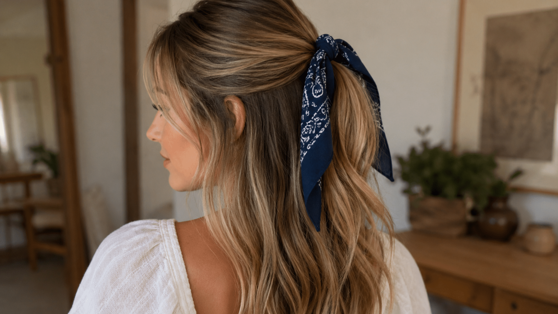 oman with long wavy blonde hair wearing a navy paisley bandana tied around a half-up hairstyle in a casual indoor setting.