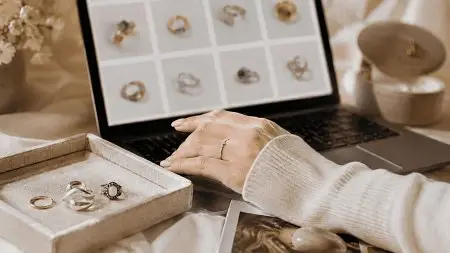 Person browsing jewelry on laptop with ring box beside, in cozy setting