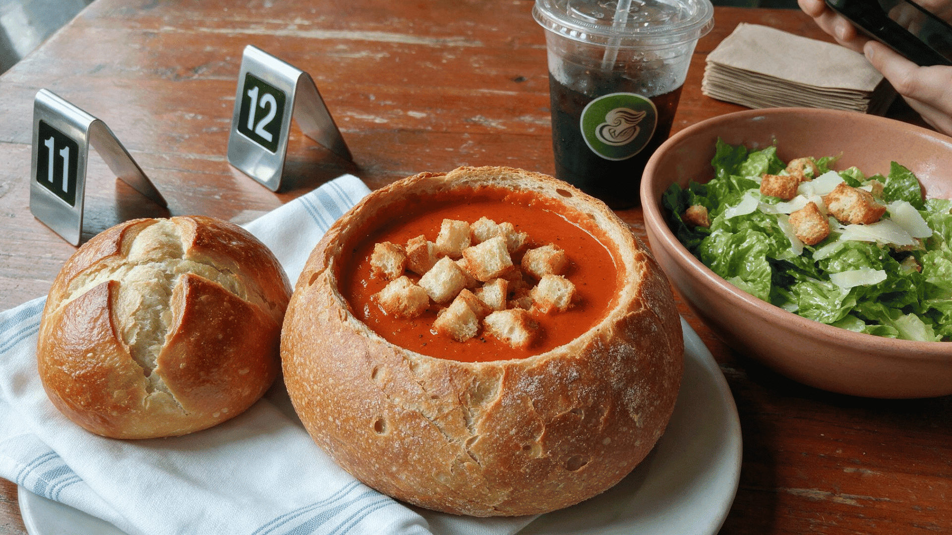 panera Bread tomato soup in a bread bowl with croutons, served beside a green salad and a cold drink.