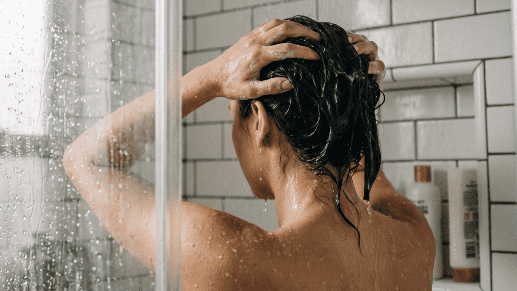 Person washing hair in a shower with wet hair and water droplets on glass.