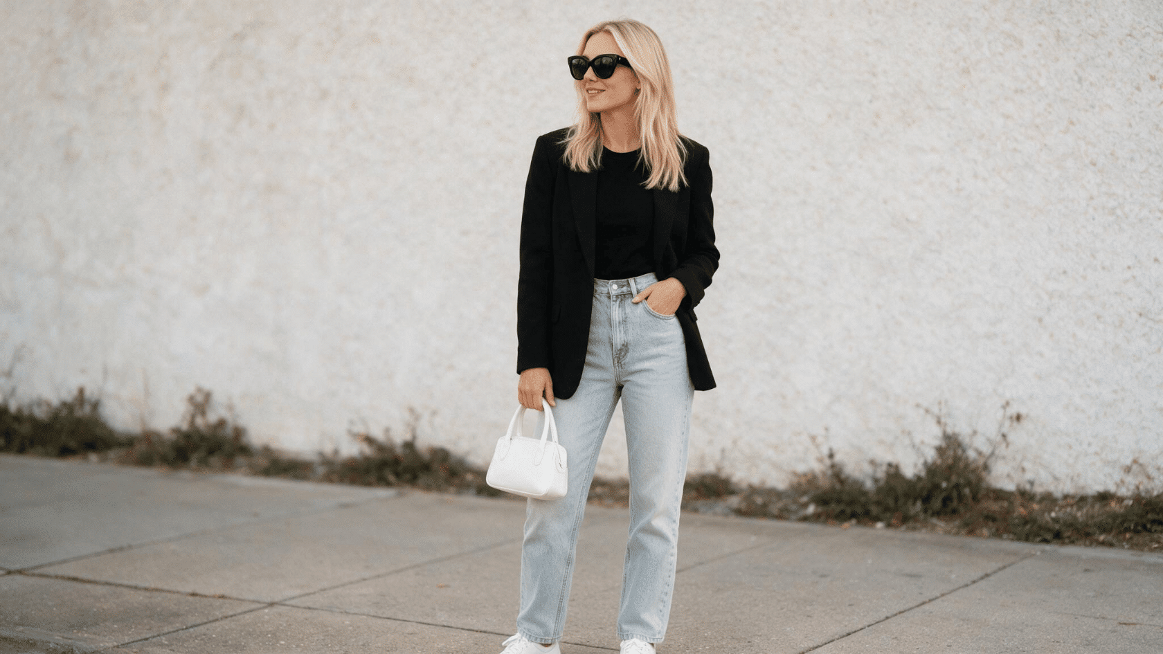 Person wearing a black blazer and light jeans with sunglasses standing outdoors in a minimalist fashion look.