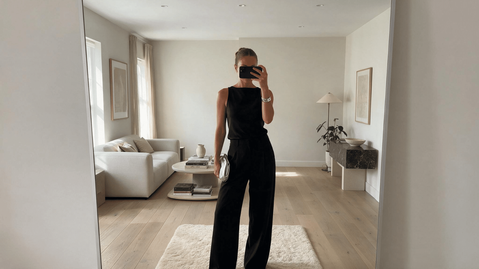 Person wearing a black outfit taking a mirror selfie in a minimalist living room.