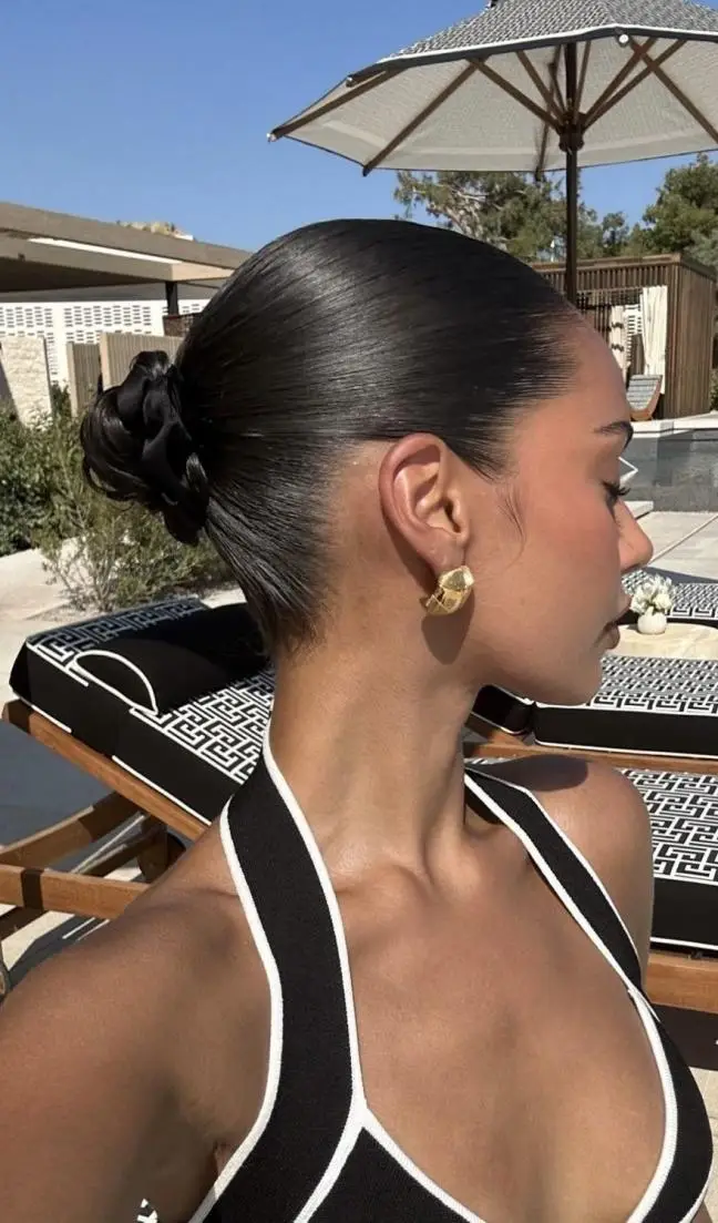 Woman with slicked-back bun wearing gold earrings beside poolside lounge area