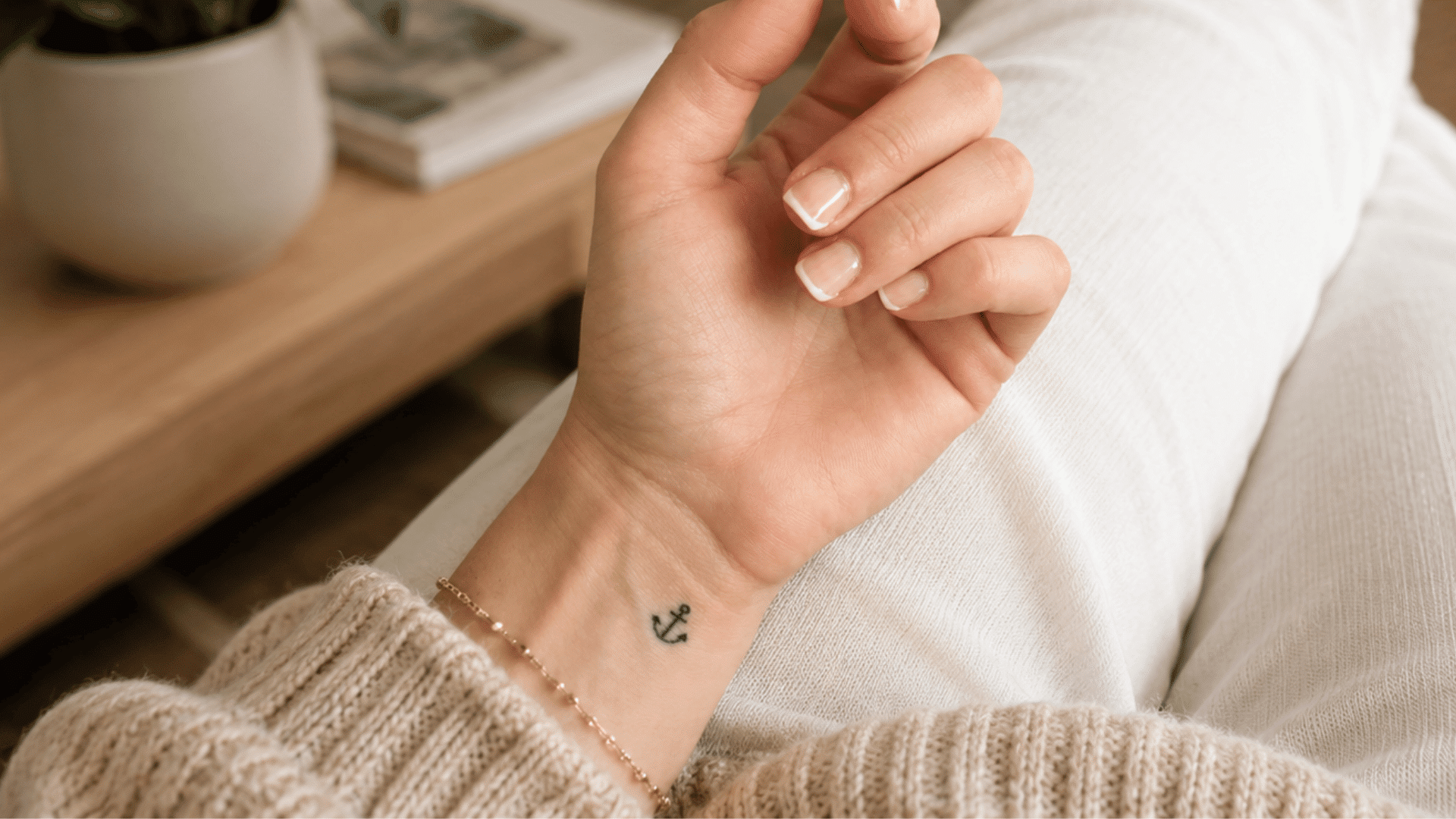 small black anchor tattoo on the wrist of a woman wearing a beige knit sweater and cream pants.