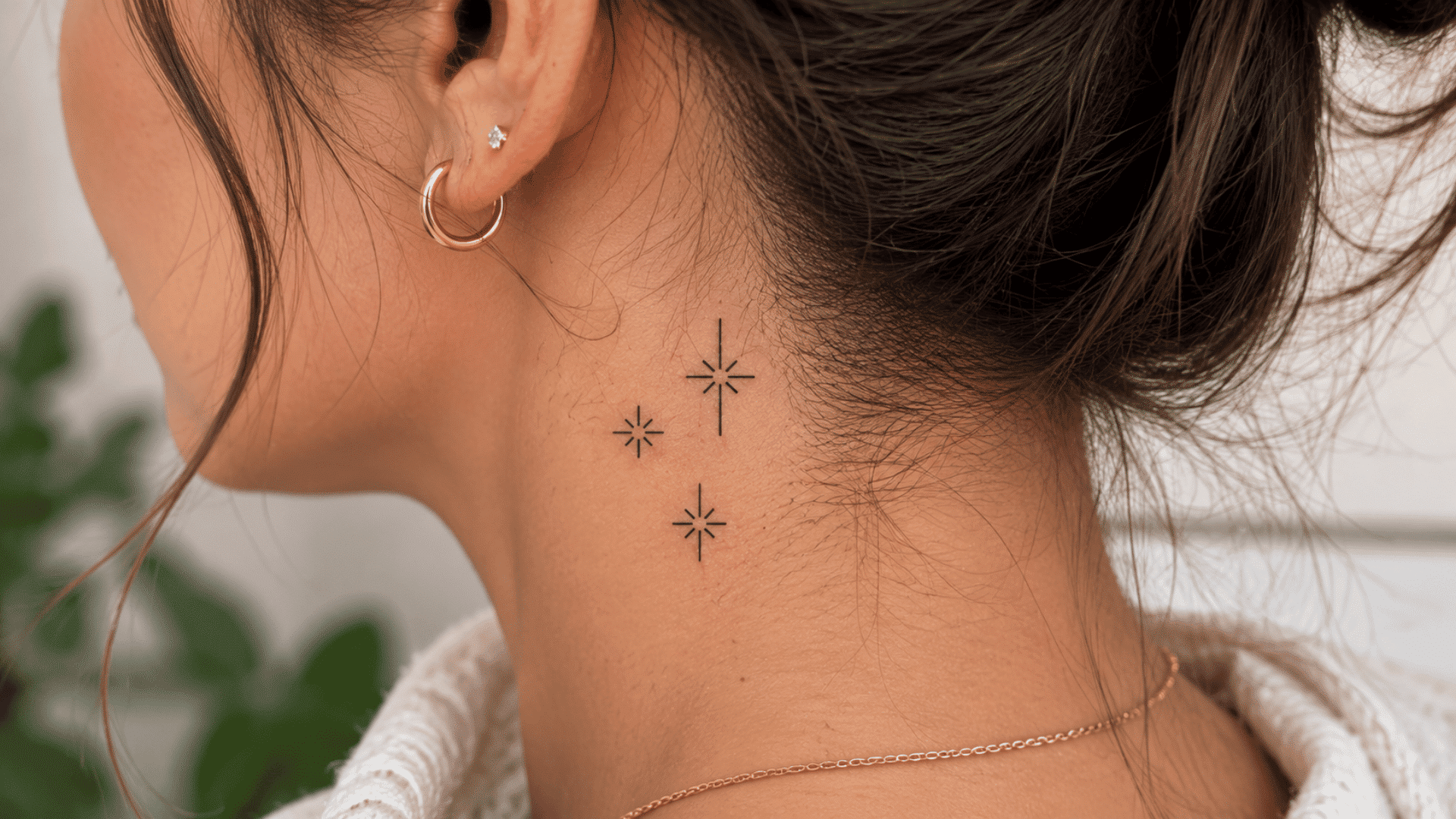 small black star tattoos behind a woman’s ear with gold earrings and dark hair.