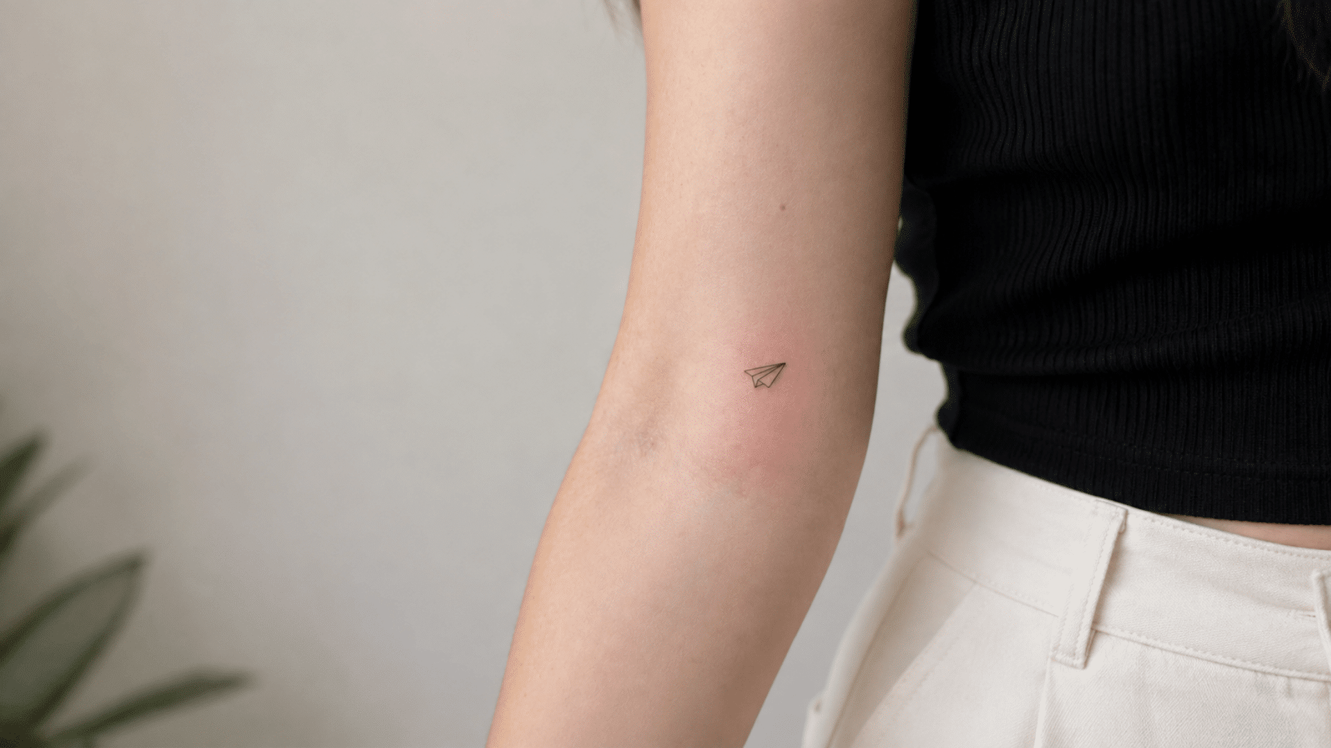 small fine-line paper airplane tattoo on a woman’s inner arm near the elbow, paired with a black top and white pants.