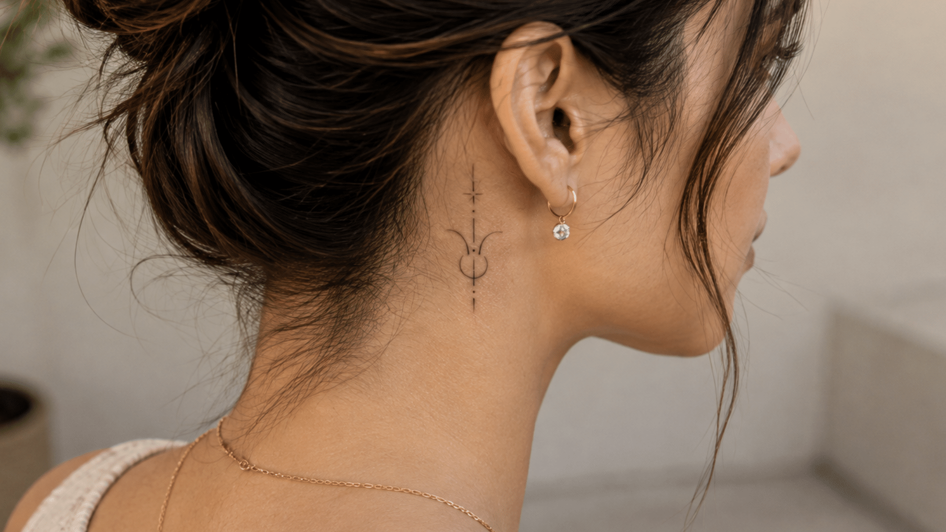 small taurus zodiac tattoo behind a woman’s ear with a gold earring and loose bun.