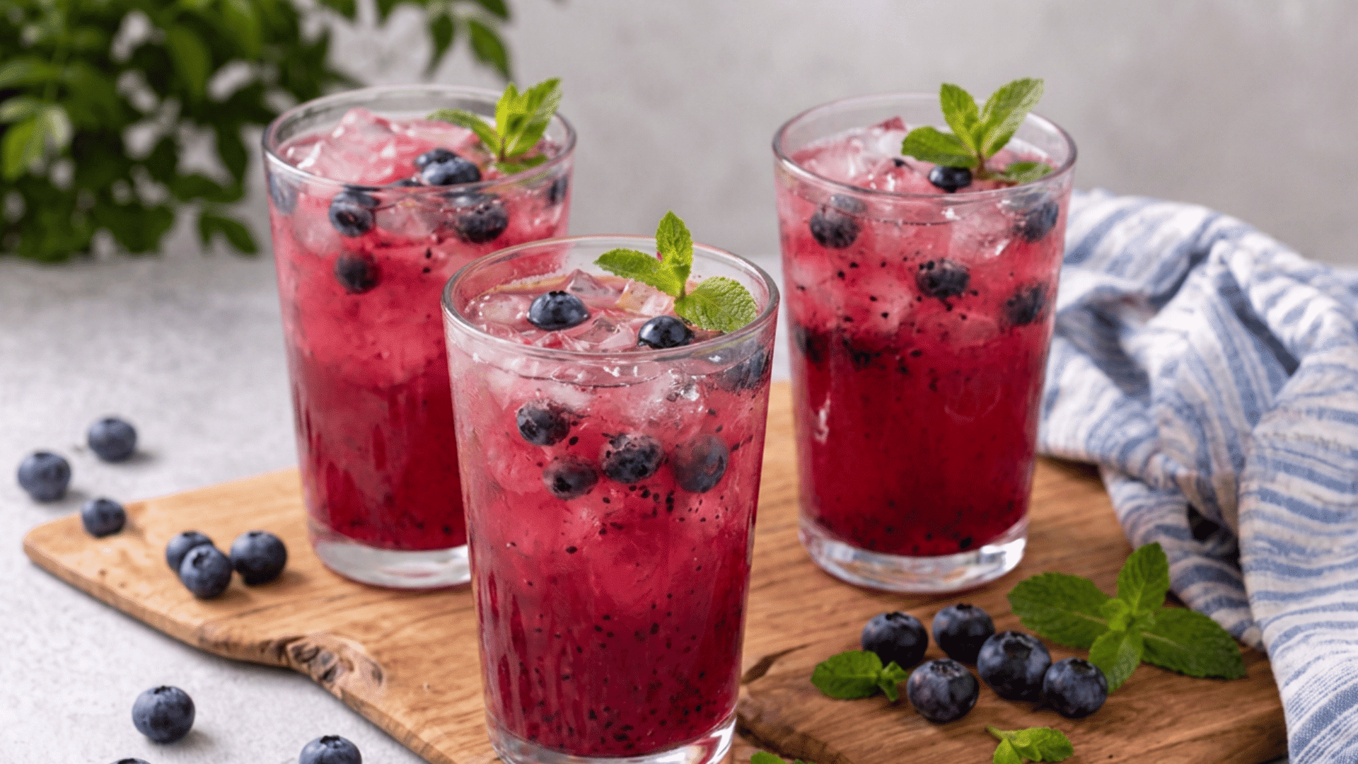 blueberry mint mocktails in glasses with ice and mint garnish, surrounded by fresh blueberries on a wooden board.