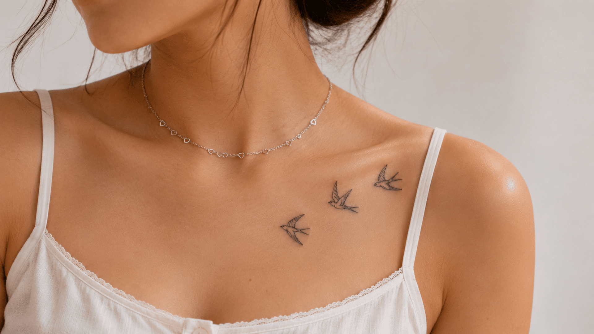 three small fine-line swallow tattoos flying across a woman’s collarbone above a white camisole.