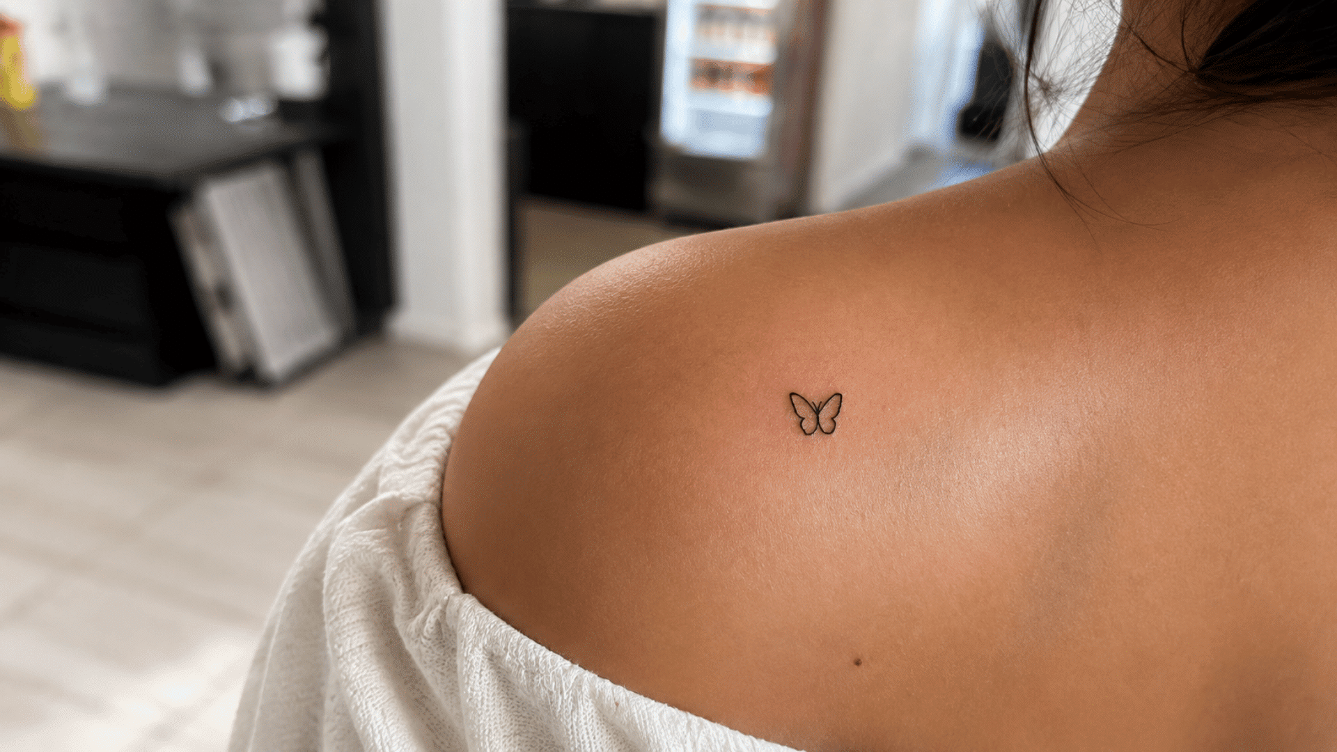 tiny minimalist butterfly tattoo on the upper shoulder with a white off-shoulder top in a softly lit indoor setting.
