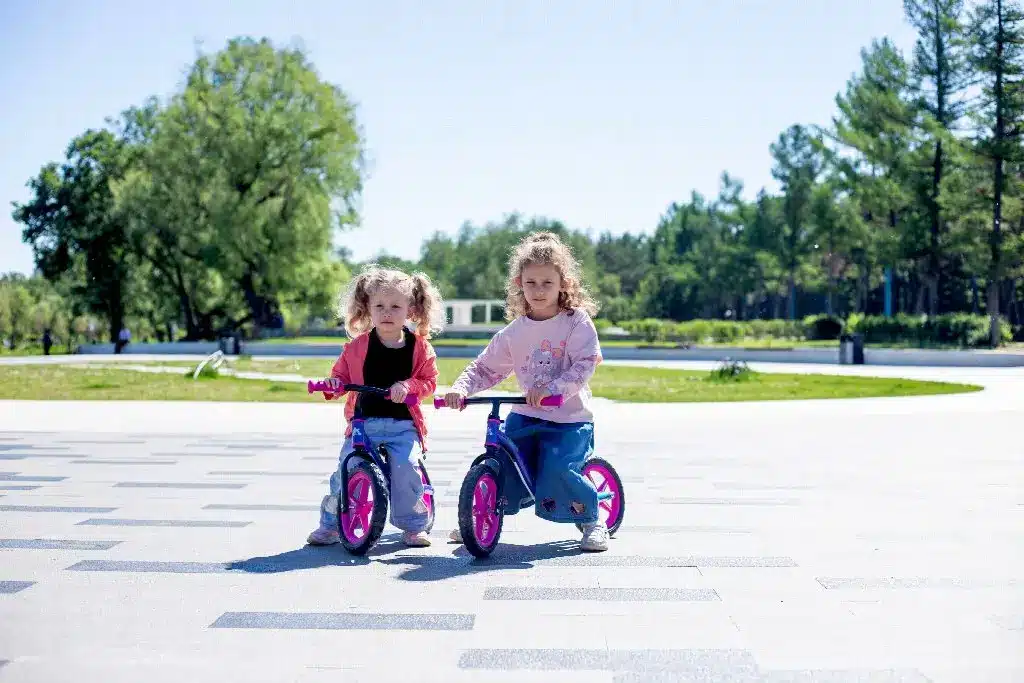 Two children riding bicycles on a sunny day in a park with trees in the background