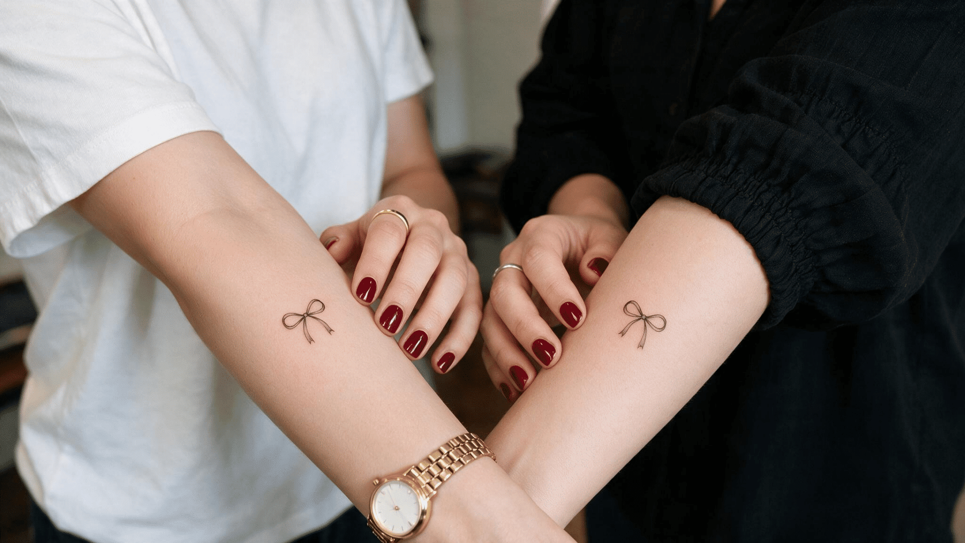 two people showing matching small bow tattoos on their inner forearms, with dark red nails and gold and silver jewelry.