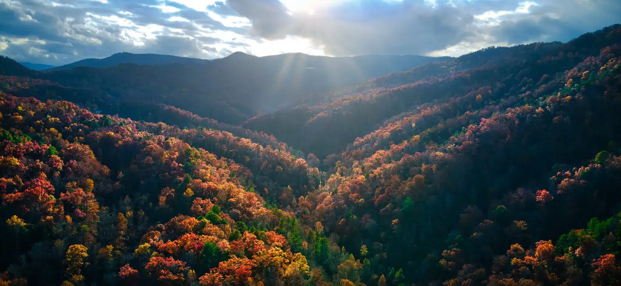 Sunlight streaming over autumn-colored forested mountains under a partly cloudy sky