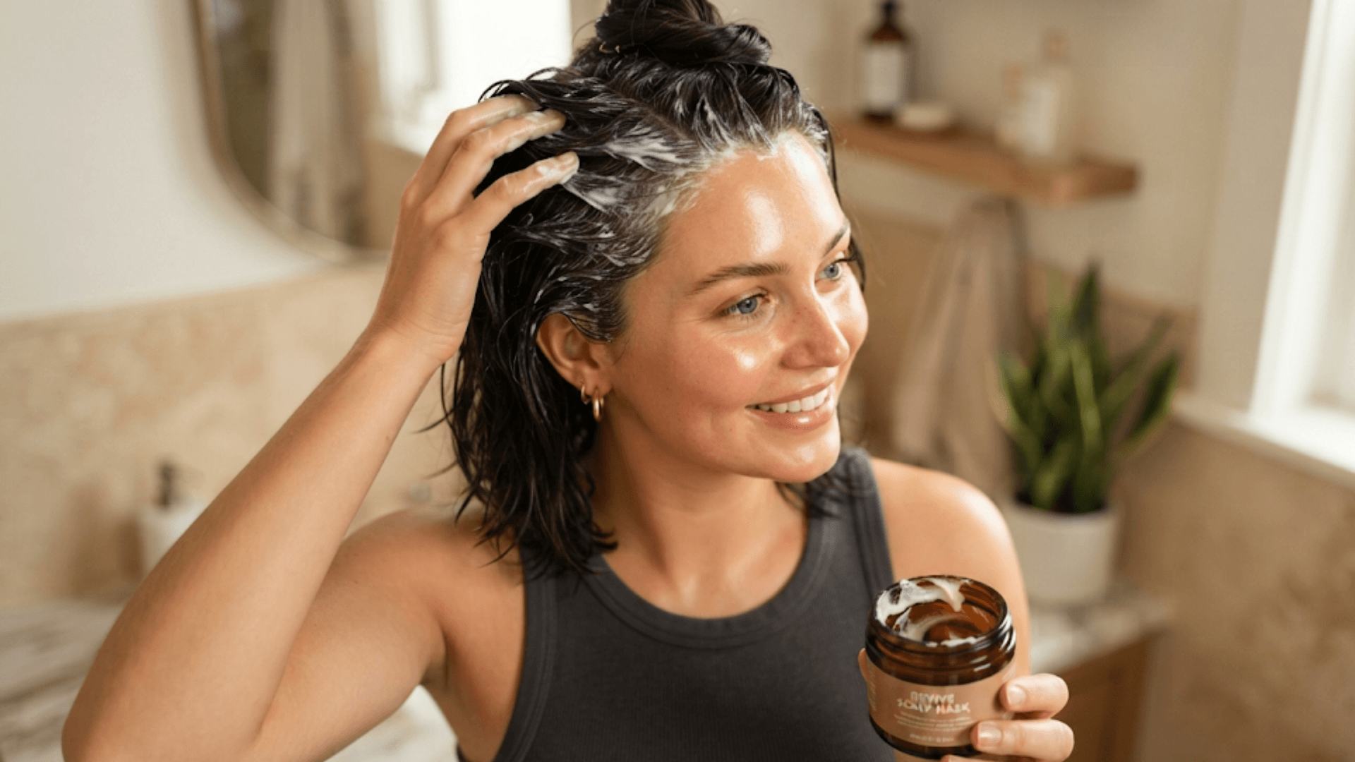 woman applying a moisturizing hair mask to wet dark hair while holding a jar in a bright bathroom