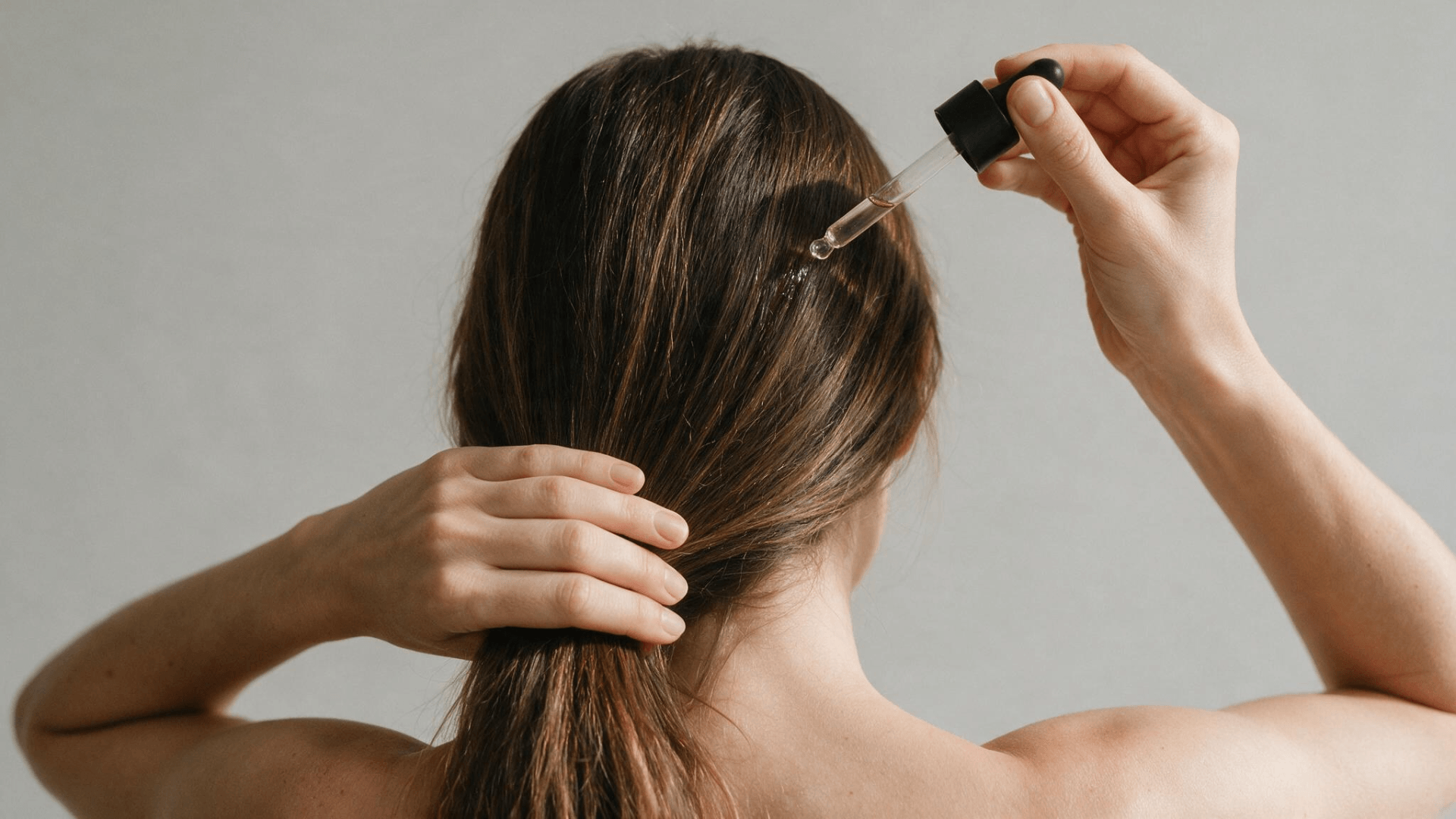 woman applying anti frizz serum to long brown hair with a glass dropper against a plain background