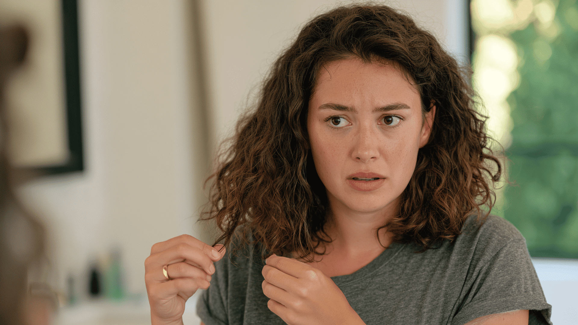 woman examining the ends of her shoulder length curly brown hair near a bright window