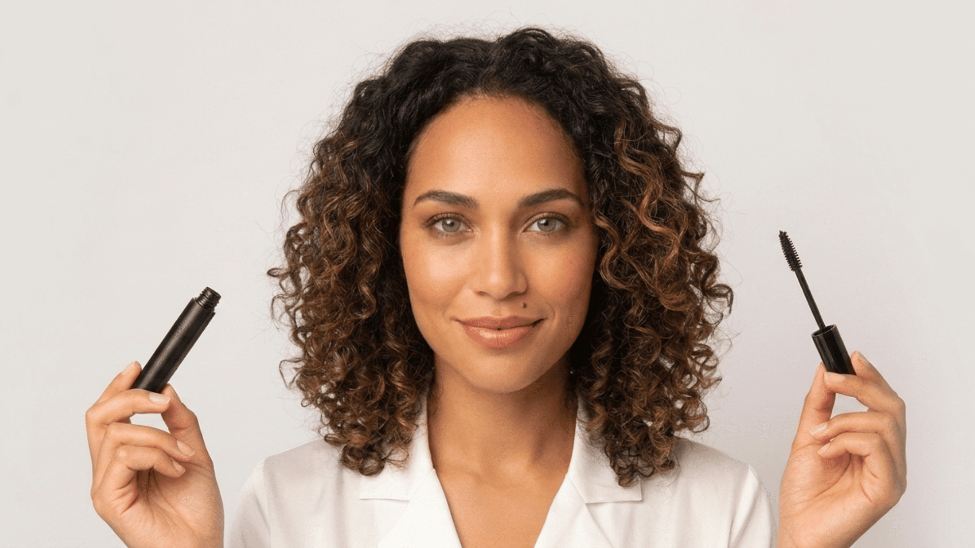 Woman holding a tubing mascara tube and wand with defined lashes against a light background.