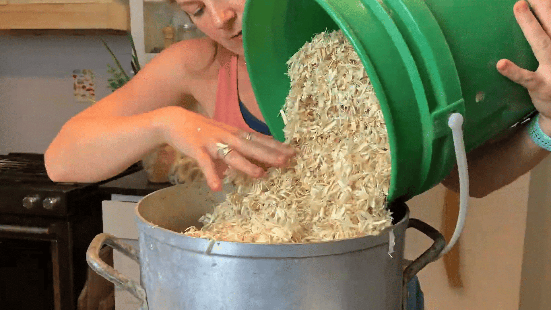 woman pouring aspen shavings from a green bucket into a large metal pot for soaking and cooling