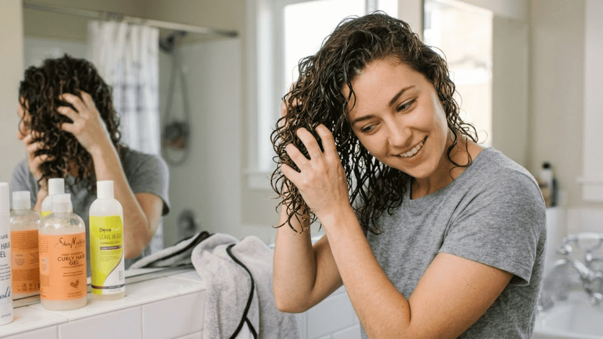 woman scrunching wet curls during her curly hair routine with styling products beside her bathroom mirror