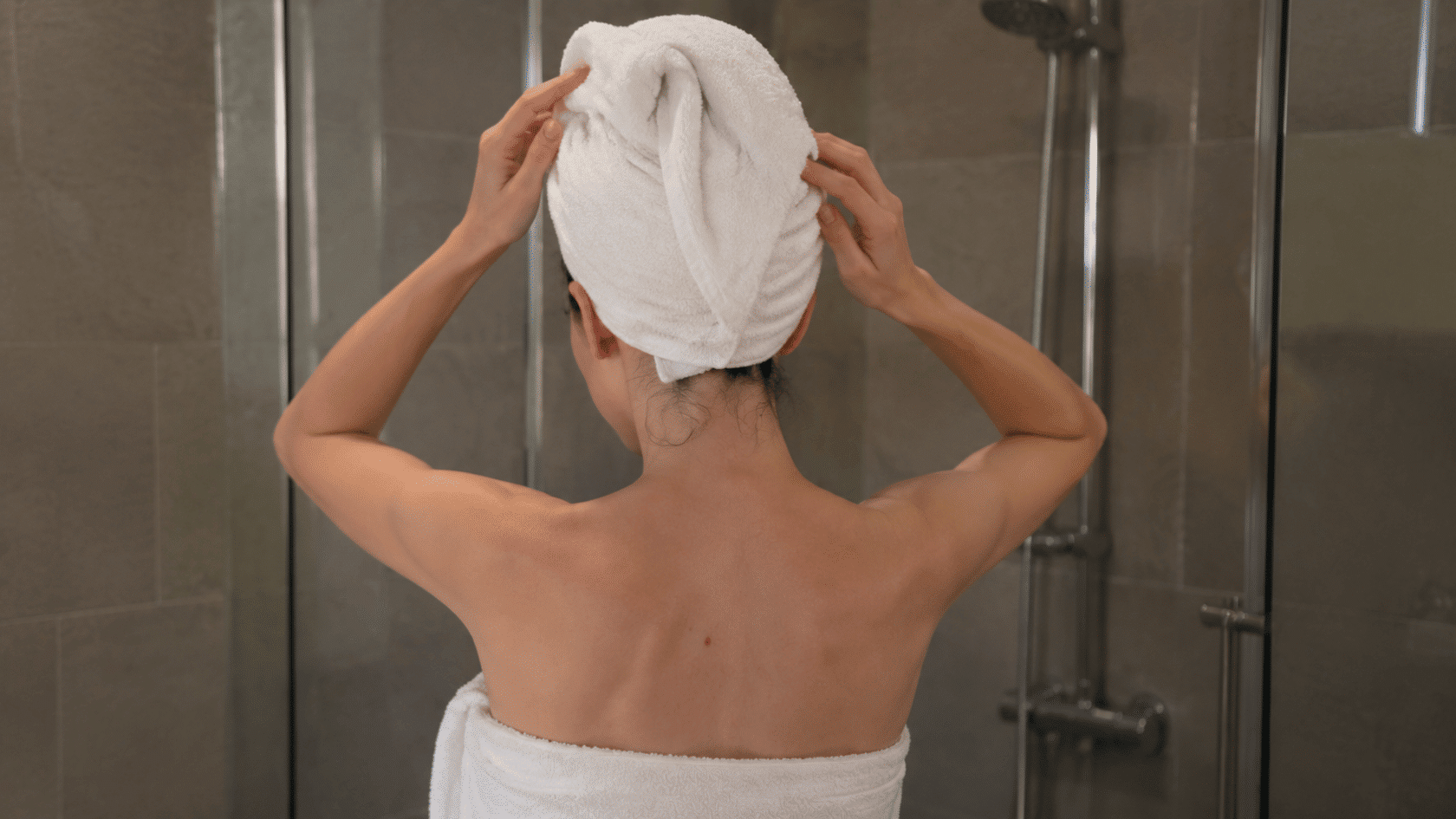 Woman standing in a shower with a towel wrapped around her hair and body, adjusting the towel while facing away from the camera.