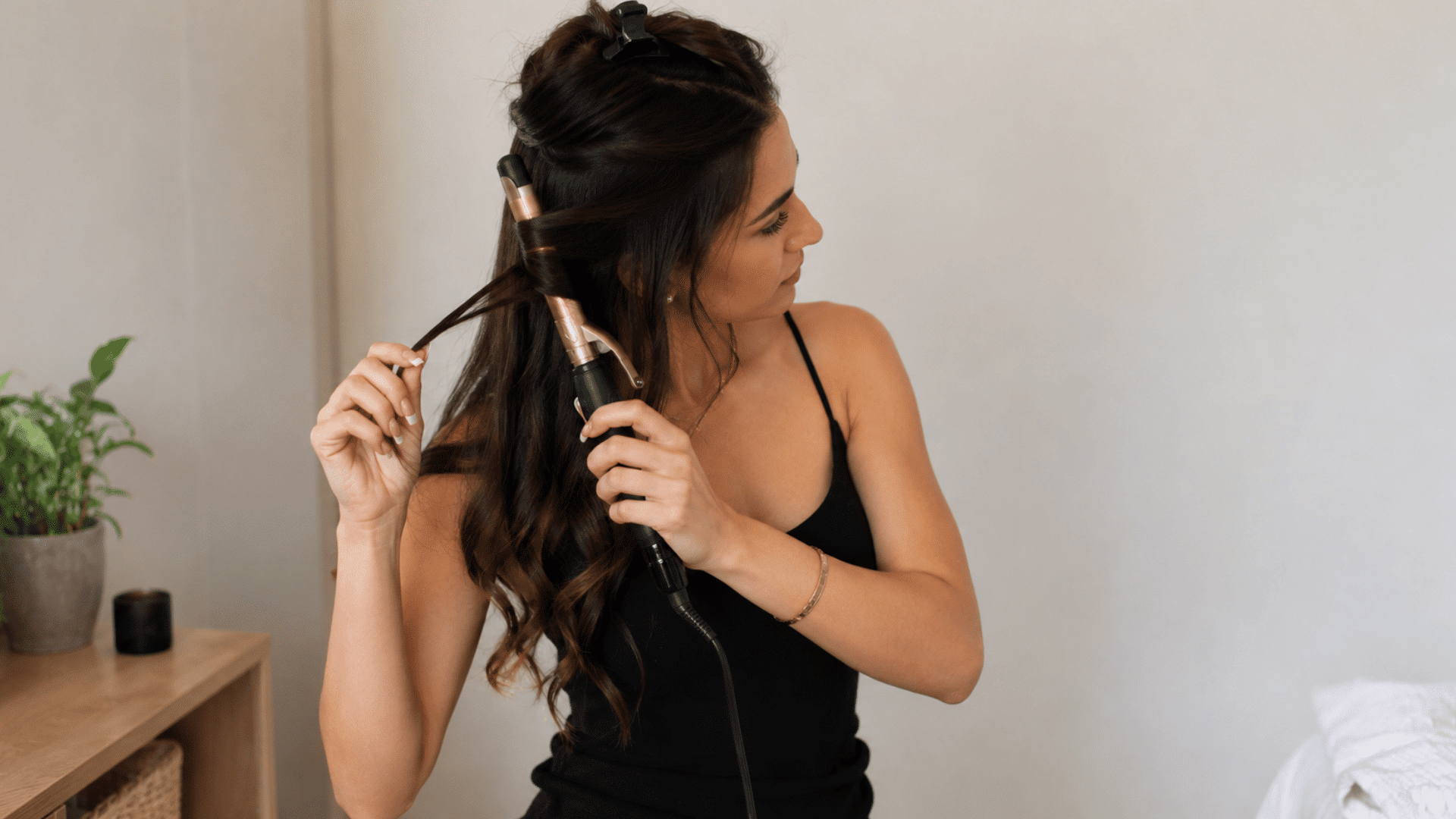 woman using a curling iron to style long hair at home in a bedroom setting