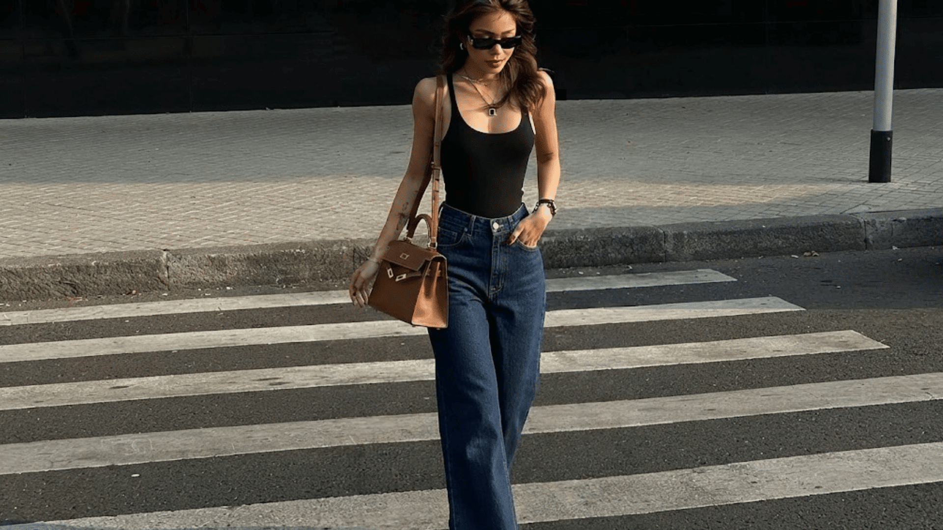woman wearing a black tank top and wide leg jeans with sunglasses and a brown shoulder bag walking outdoors