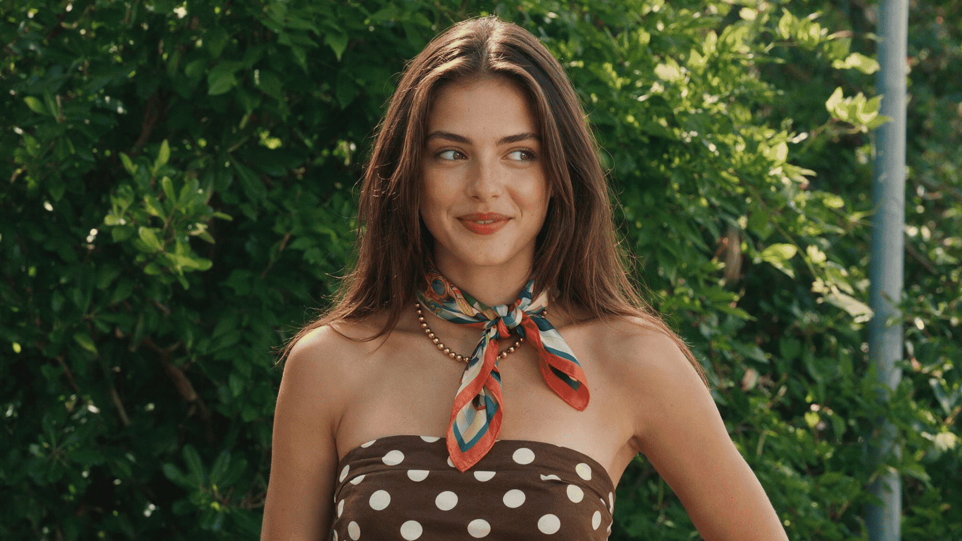 woman wearing a colorful bandana tied around her neck with a brown polka-dot strapless top, standing outdoors against a leafy green background.