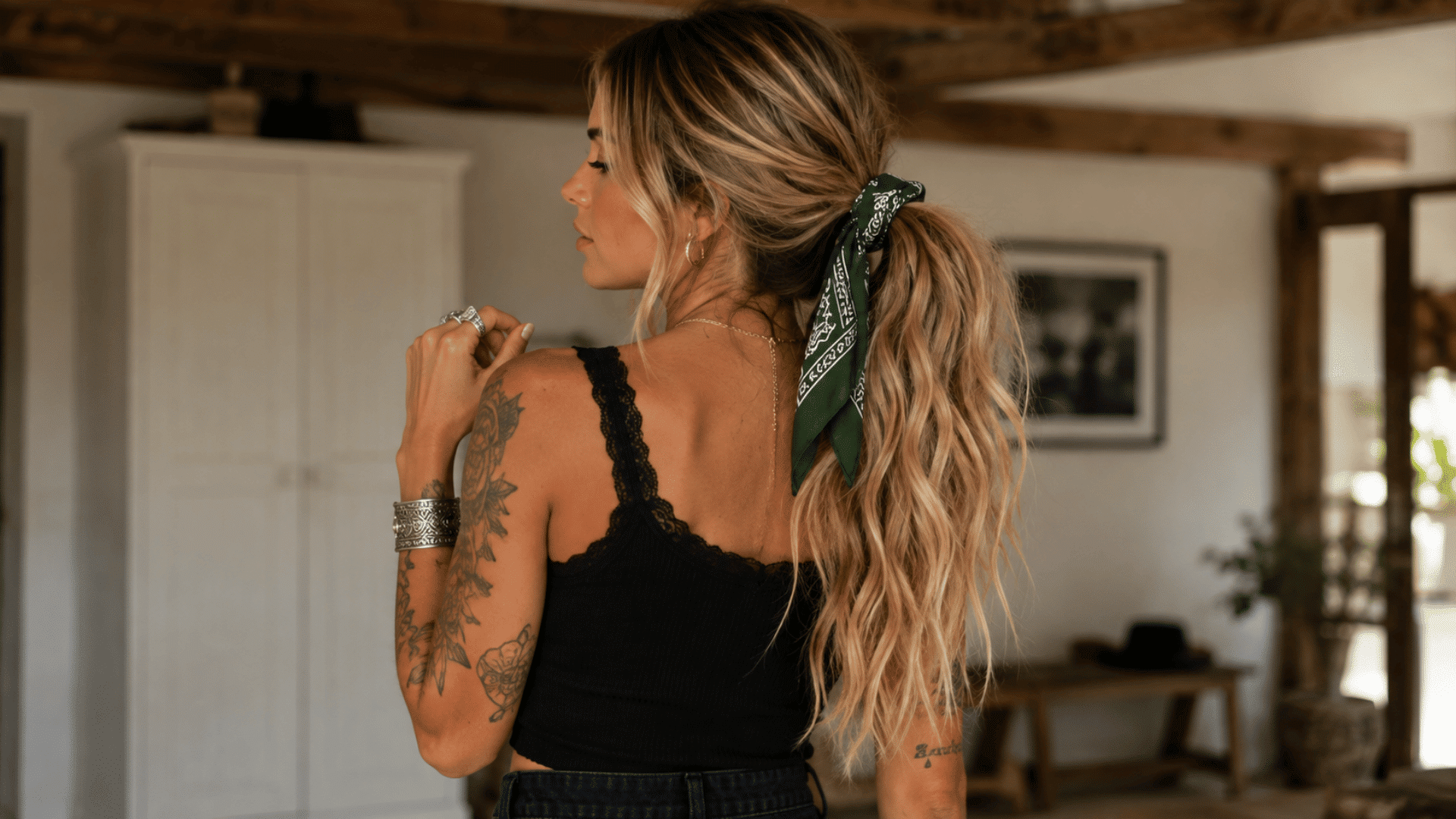 woman with long blonde wavy hair wearing a green paisley bandana tied around a ponytail, paired with a black lace-trim tank top.