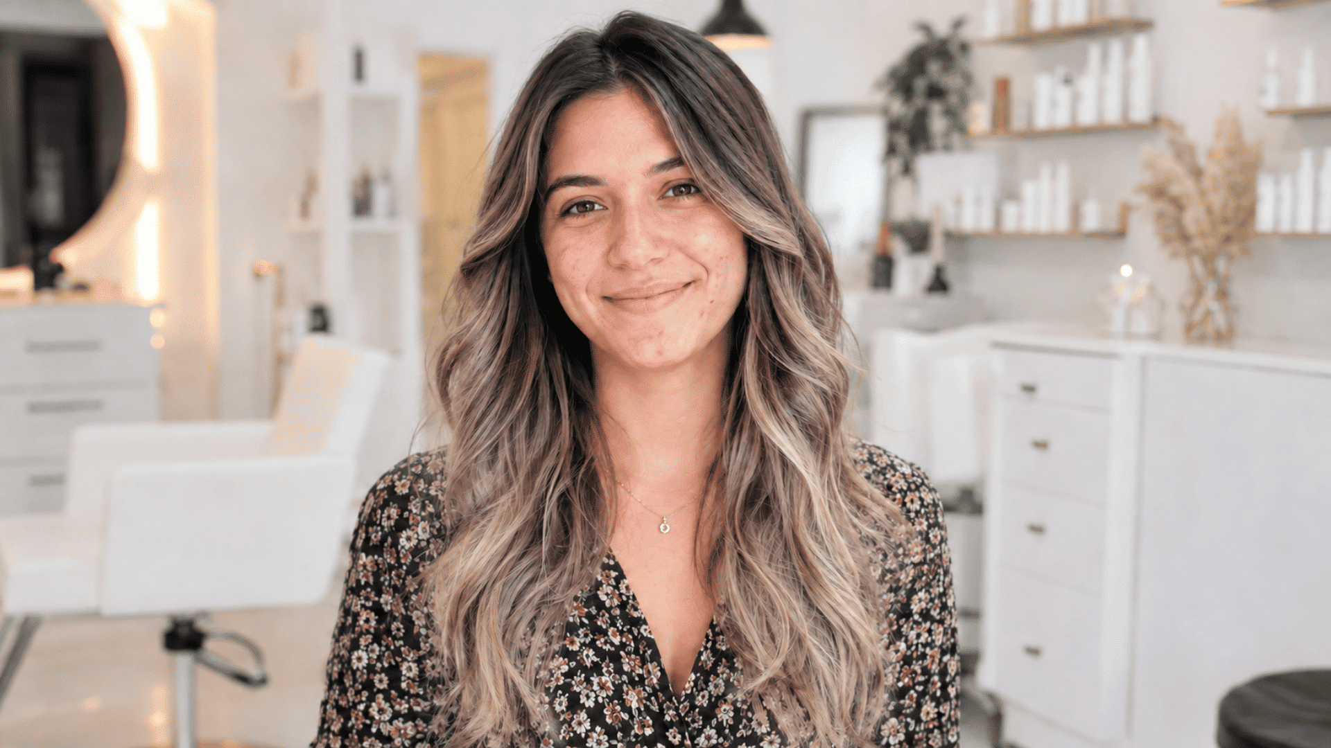woman with long wavy dark brown hair and beige blonde money piece highlights smiling in a bright salon