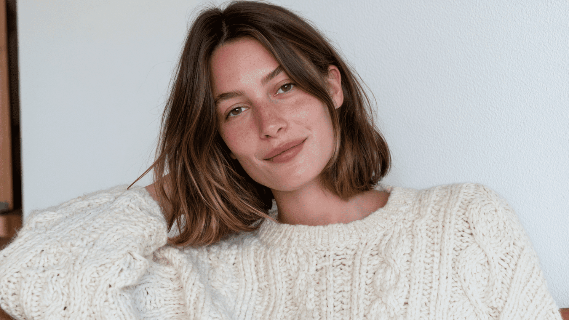 woman with thick shoulder length brown hair wearing a cream sweater against a light wall