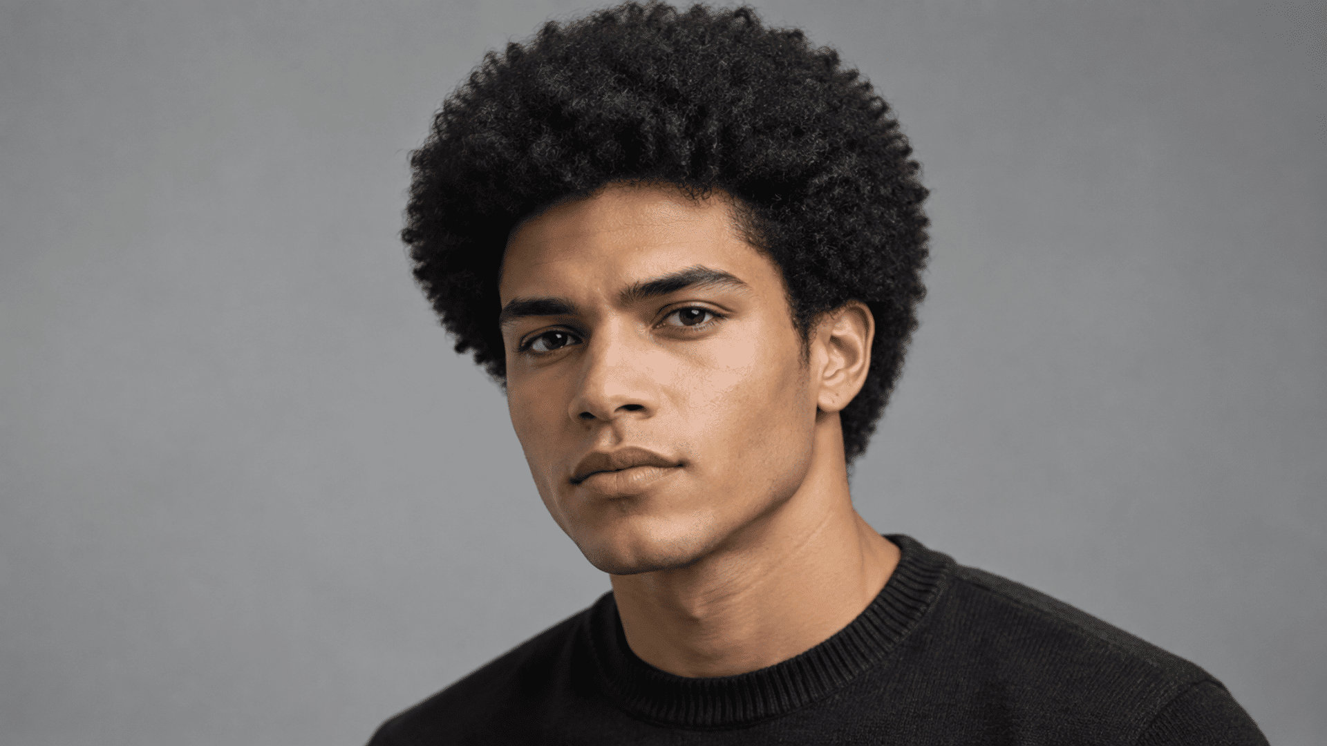 young man with curly afro hairstyle wearing black sweater against a neutral gray background portrait