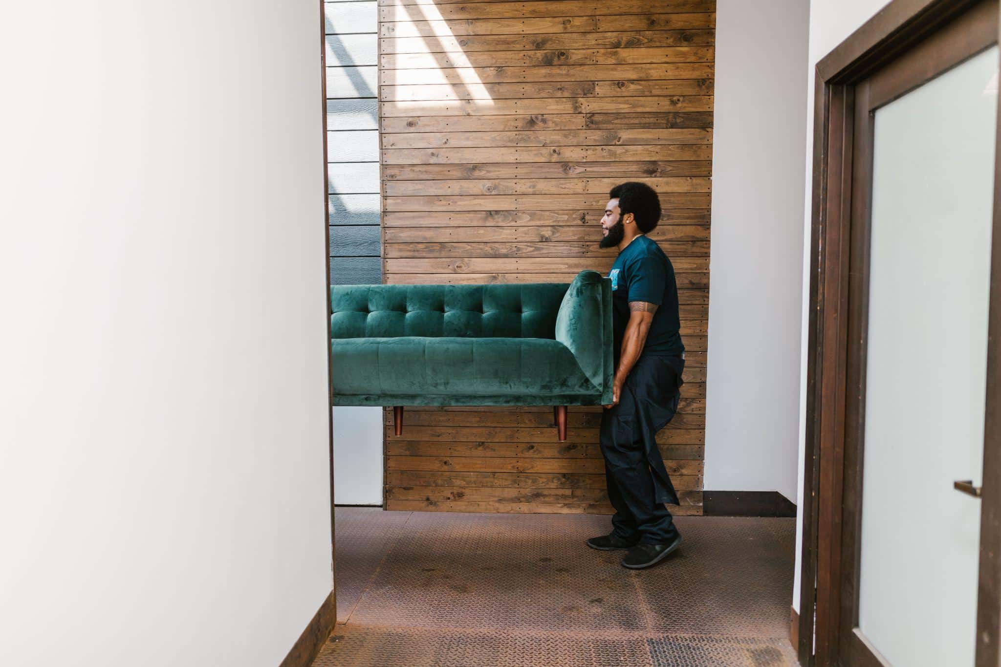 Man carrying a teal velvet sofa in a modern, wooden-accented hallway