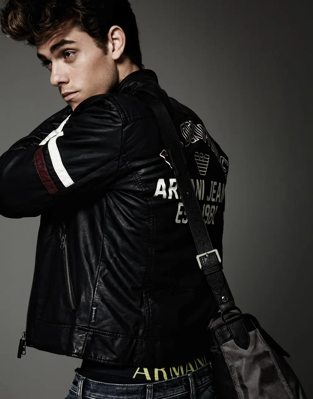 Young man in black leather jacket with shoulder bag in moody studio setting