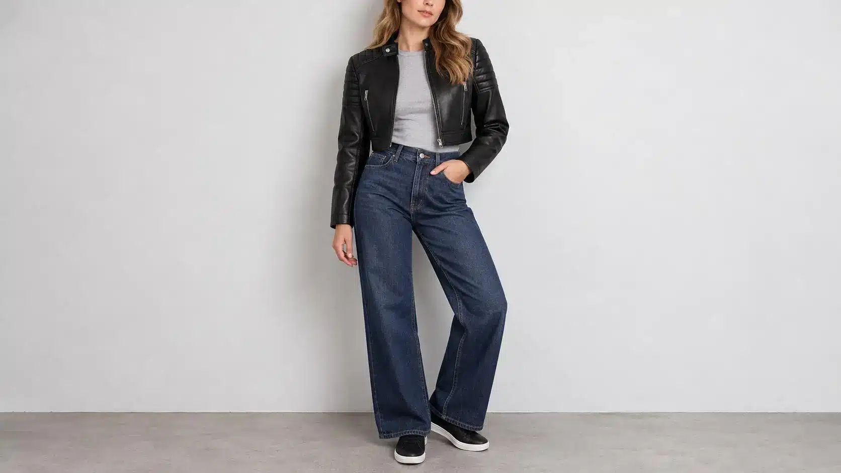 Woman wearing black leather jacket and blue jeans standing against light gray wall