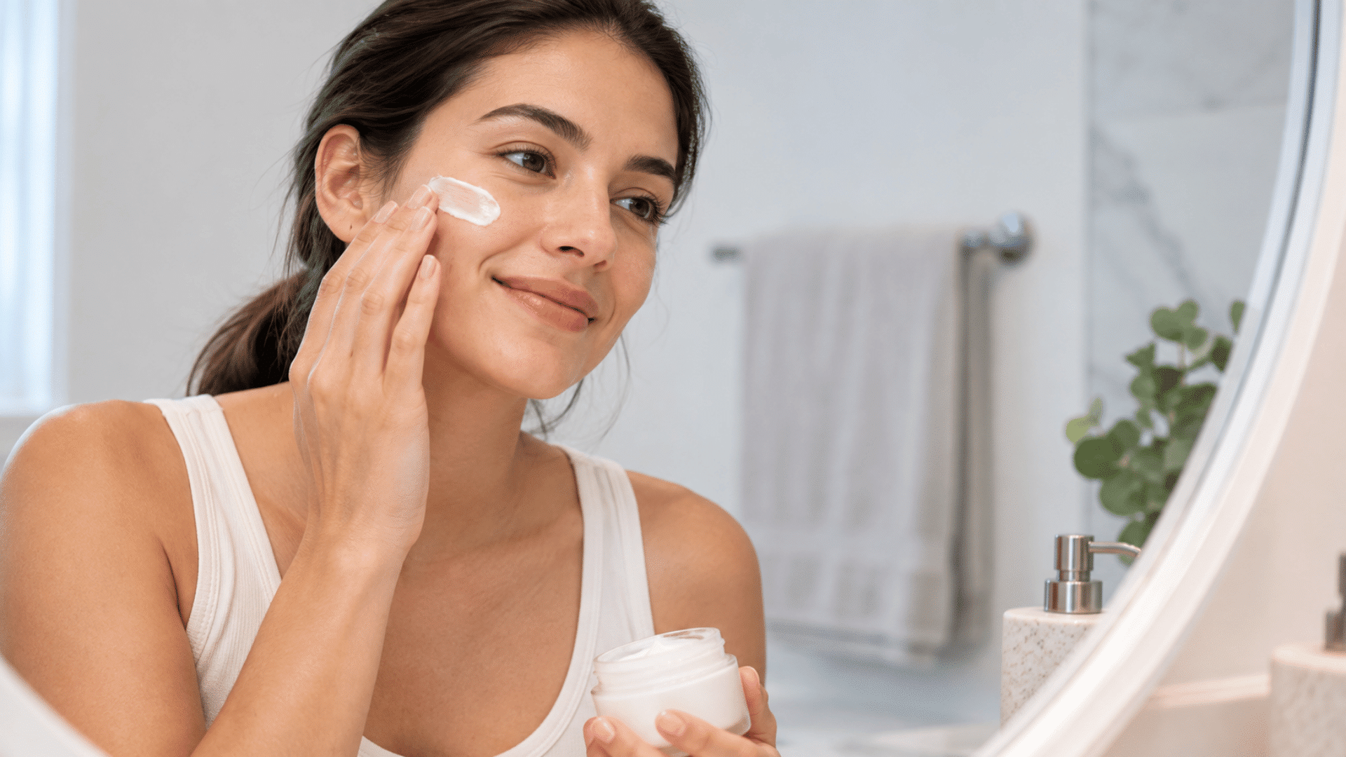 woman applying ceramide moisturizer to her cheek while holding an open cream jar in front of a mirror
