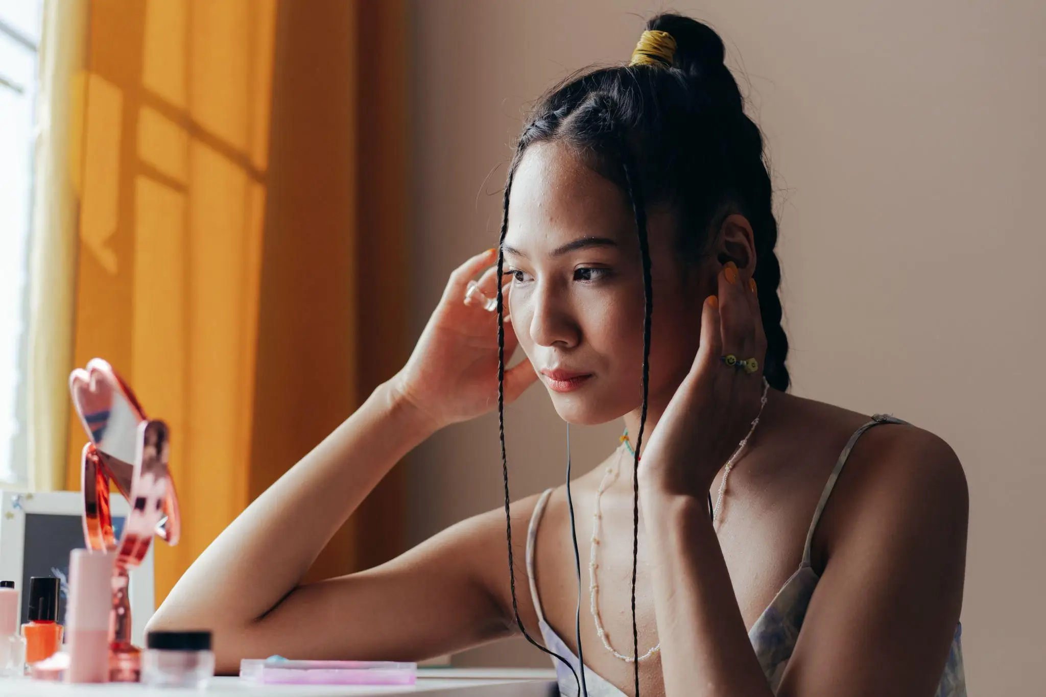 Young woman with braided hair adjusting earbuds in front of a mirror with cosmetics nearby