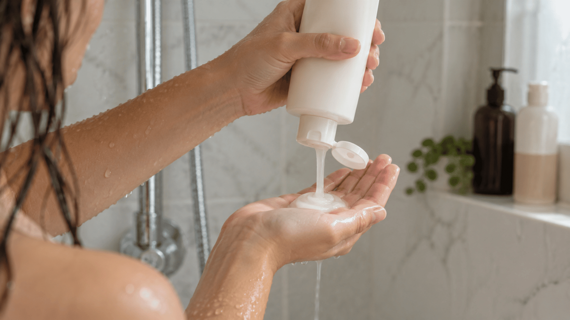 a woman pouring shampoo from a white bottle into her hand while standing under a shower with wet hair
