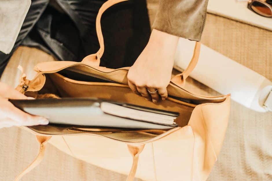 Person placing a laptop into a tan leather bag on a textured surface