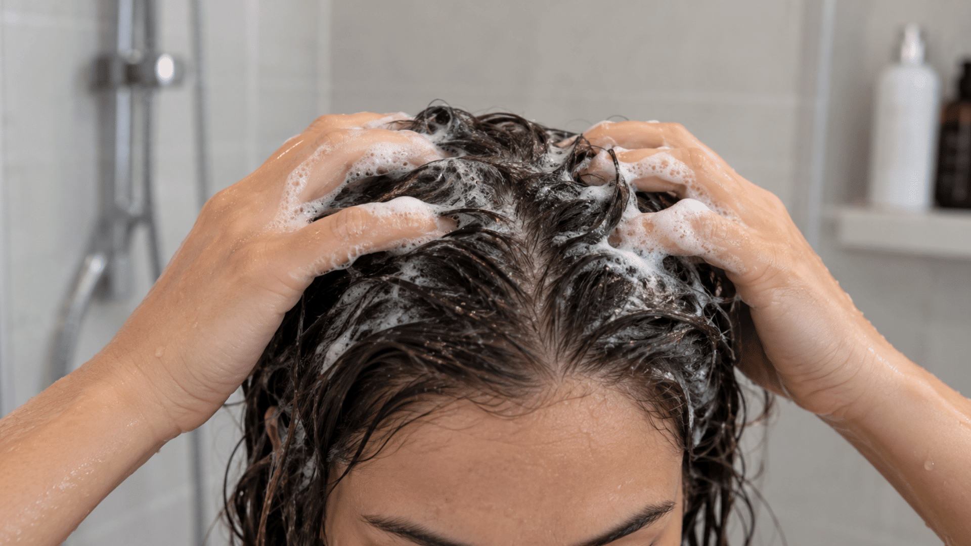 a person washing their dark wet hair with shampoo while massaging thick foamy lather into the scalp during a shower