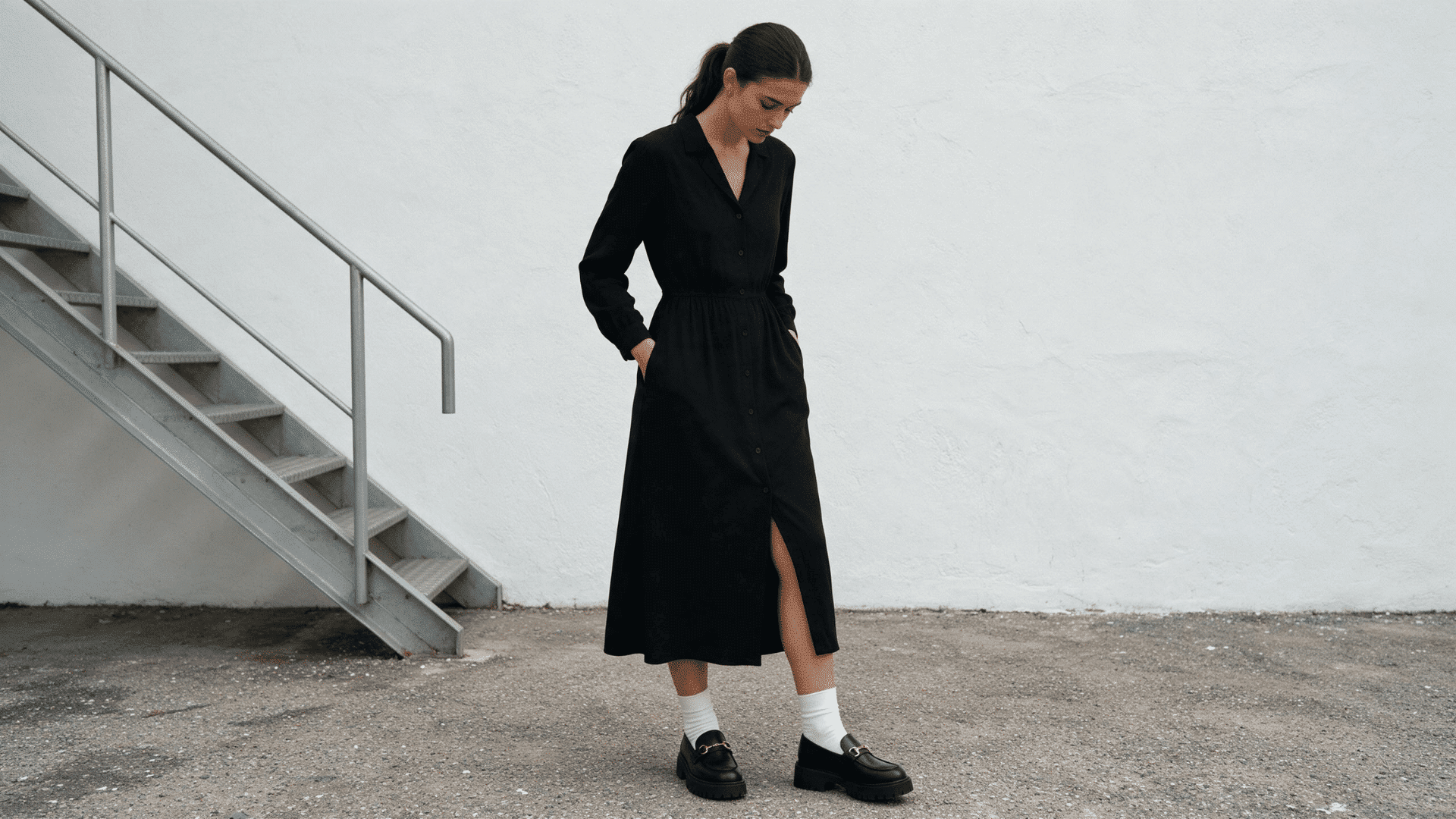 woman wearing a black midi dress with white socks and chunky black loafers near an industrial staircase