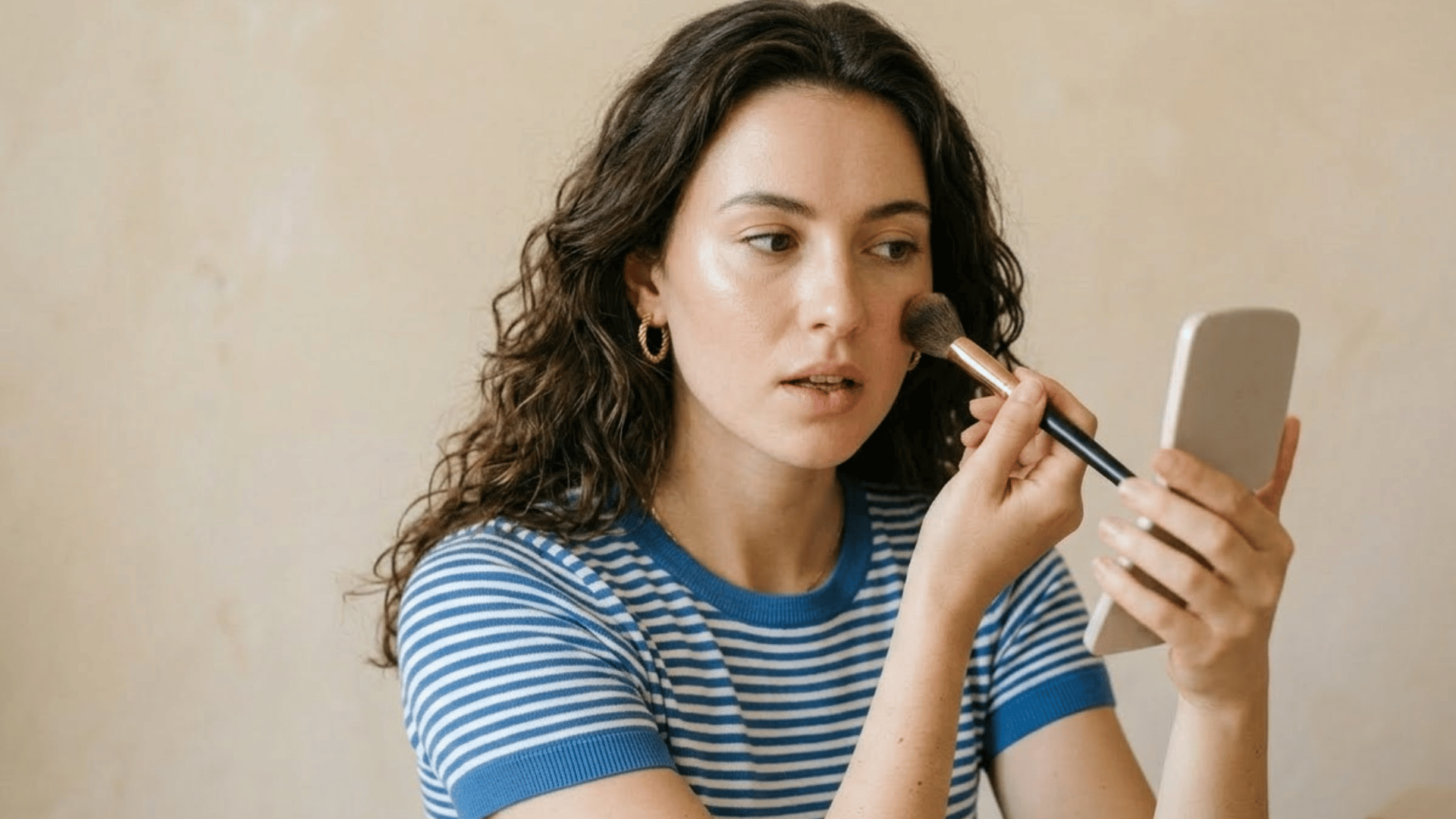 woman applying blush with a makeup brush while looking into a mirror for a soft natural everyday makeup look