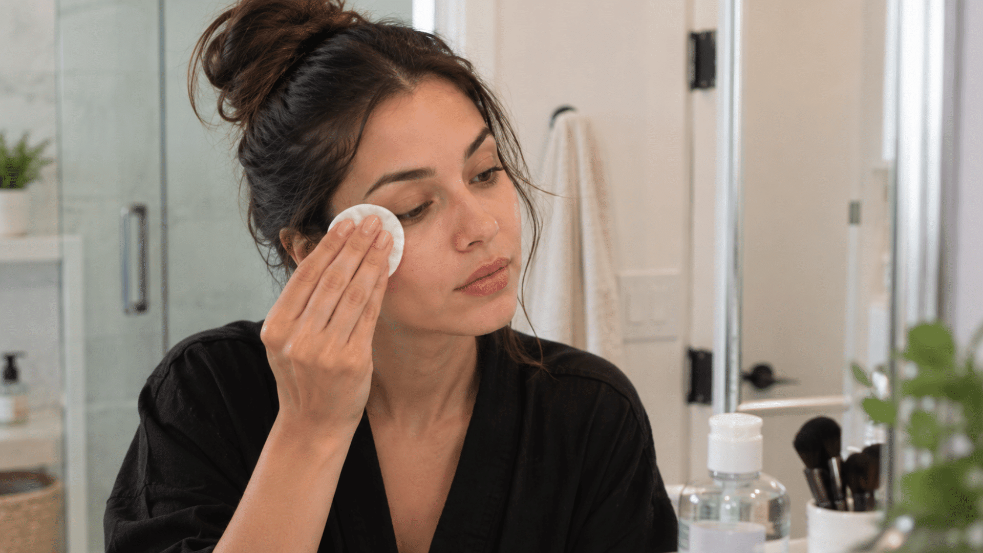 a woman removing makeup from her face with a cotton pad while looking in a bathroom mirror
