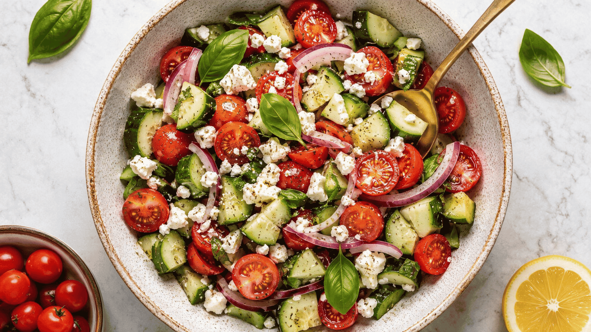 overhead view of Greek salad with cucumbers, cherry tomatoes, red onion, feta cheese, basil, and gold spoon