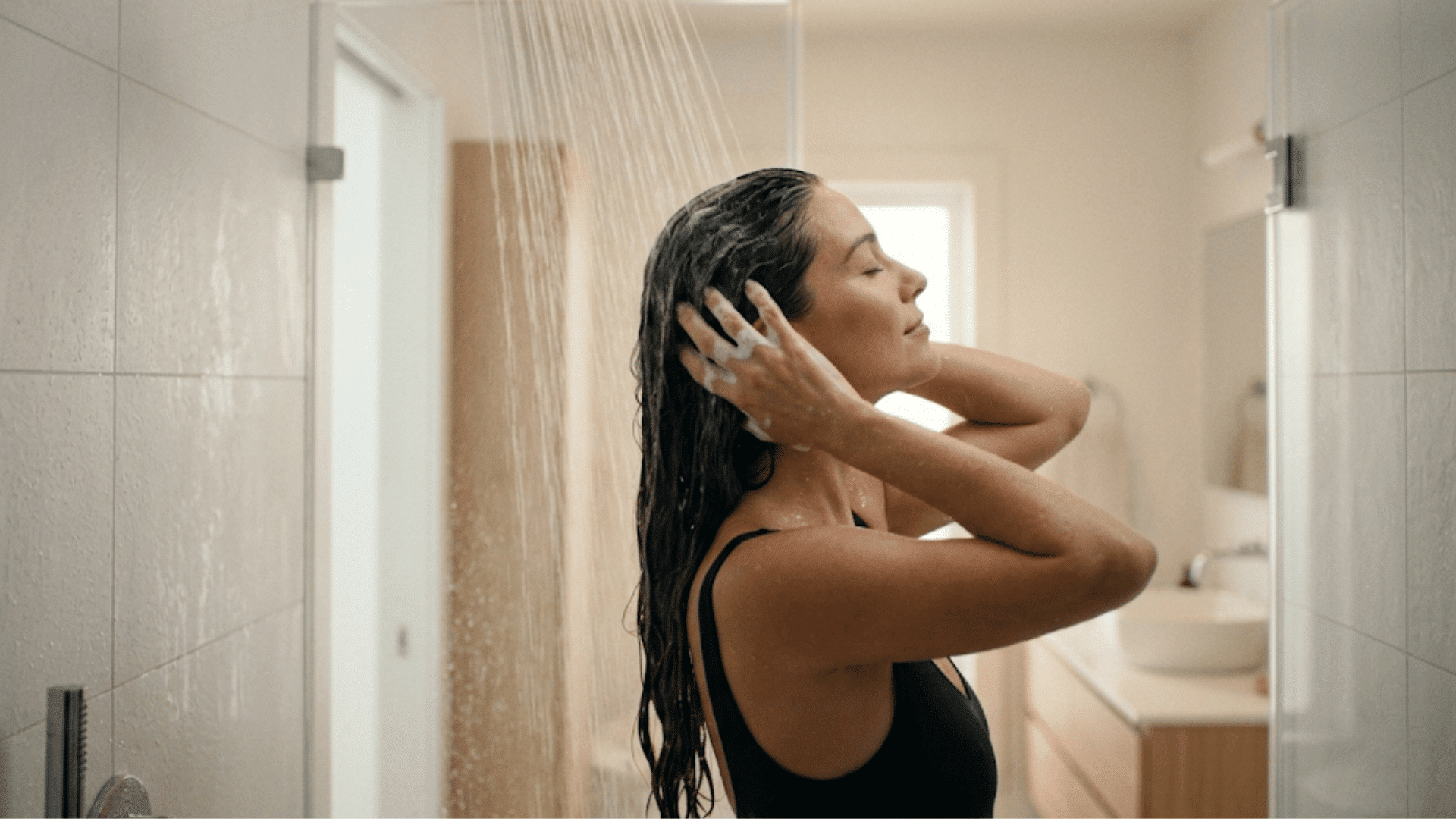 a woman washing her long dark hair under a rainfall shower while standing inside a bright modern bathroom
