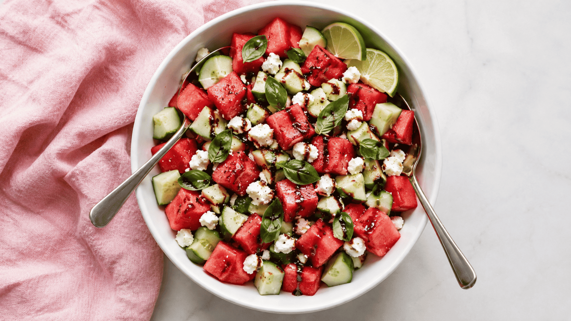 watermelon feta salad with cucumber pieces fresh basil balsamic drizzle and lime wedges in a white bowl