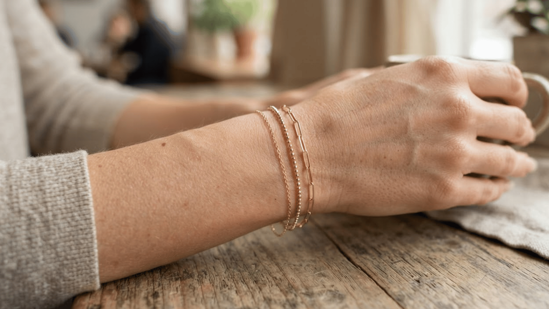 a hand wearing three delicate gold bracelets resting on a rustic wooden table in a softly lit indoor setting