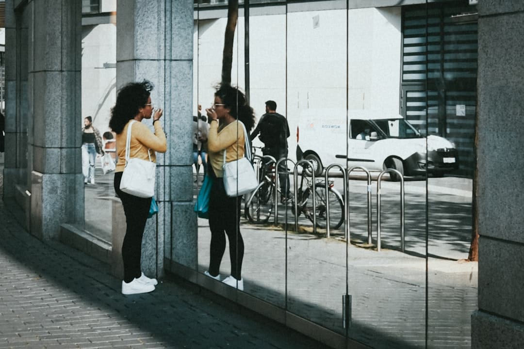 Person in yellow sweater applying lipstick reflected in mirrored building on urban sidewalk