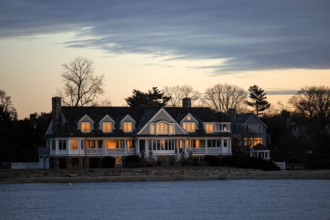 Large waterfront house with illuminated windows at dusk surrounded by bare trees